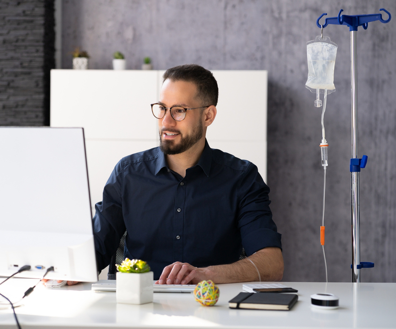 Das Foto zeigt einen Mann, der gerade Computer arbeitet und währenddessen eine Infusion erhält.